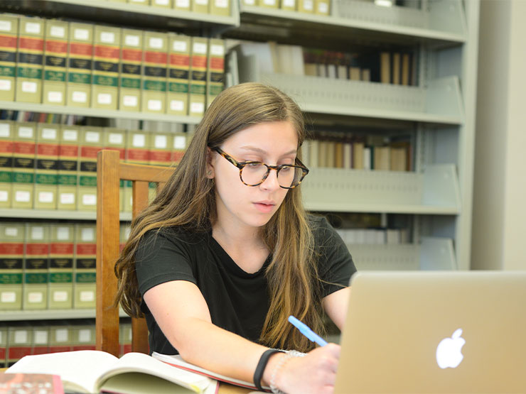 A student sits at a desk in a library writing in a notebook with a laptop open nearby.