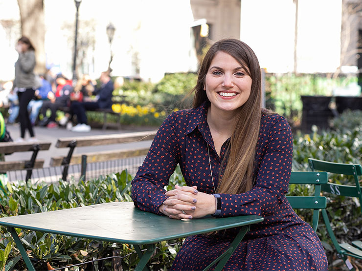 An adult with long brown hair and wearing a blue and red dress, smiles while seated a green table outdoors.