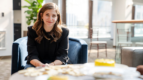 A woman with shoulder-length brown hair wears a black blazer and blouse and looks to the camera from across a table of desserts.