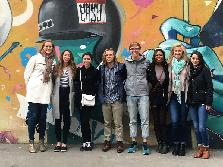 A group of students pose, smiling, with an instructor against a wall mural in a foreign country.