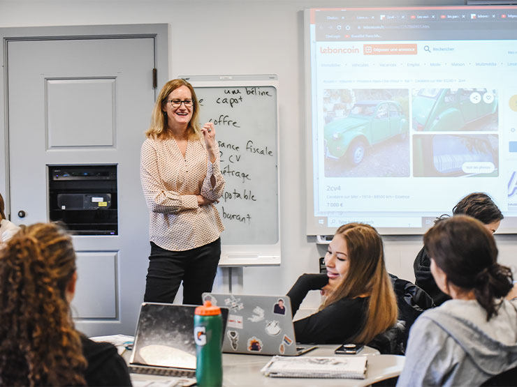 An instructor stands in front of a classroom, interacting with the class, with French words written on a white board.