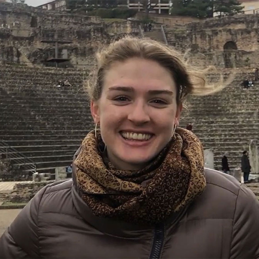 A young adult wearing a brown jacket and scarf smiles while standing in the ruins of a historic structure.