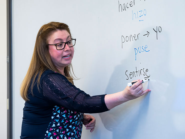 An adult with long brown hair, glasses and a blue blouse, writes Spanish words on a white board in a classroom.