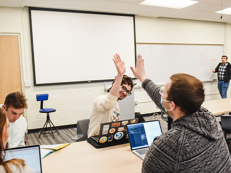 A pair of young adults high five over a table in a classroom.