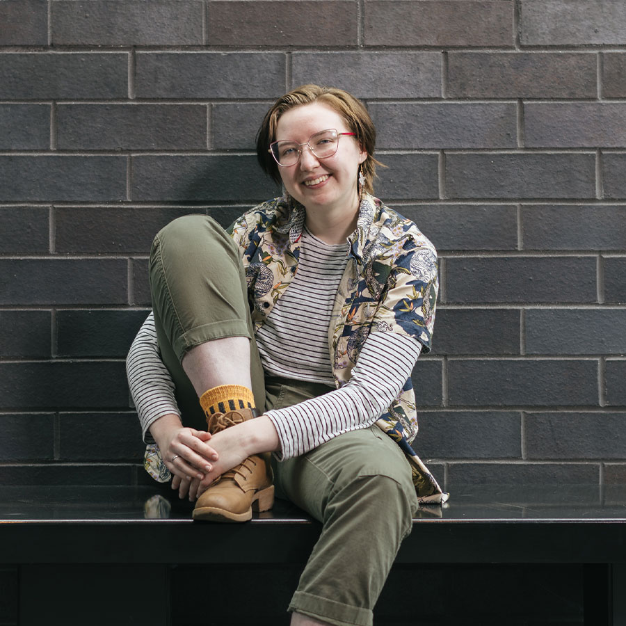 A young adult with short hair and glasses smiles and sits with a knee propped up on a bench against a grey brick wall.