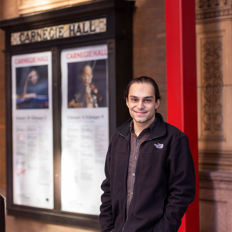 A smiling adult in a collared shirt and black jacket stands outside of a building with the sign 