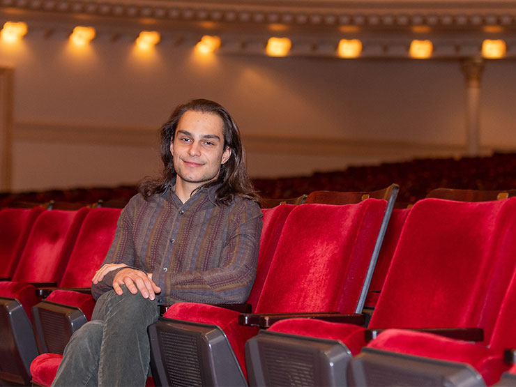 An adult with shoulder-length hair sits in an empty concert hall filled with red velvet seats.