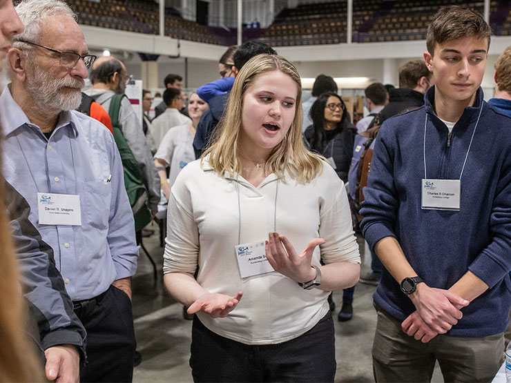 A young adult speaks about a poster presentation to a group of people standing nearby, all wearing event nametags around their necks.