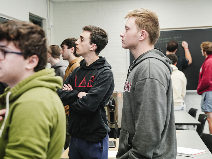 College students in a mathematics class stand in front of and contemplate problems on a chalkboard.