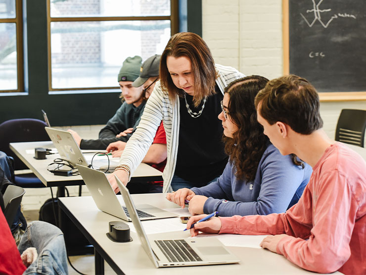 An instructor leans over the shoulder of a pair of students and points to the screen of an open laptop.