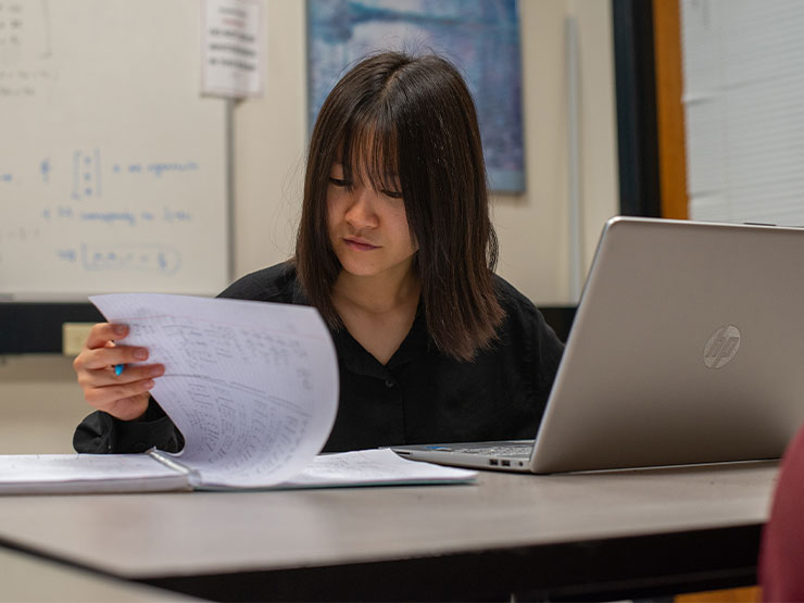 A student works on a laptop and leafs through documents in a classroom.