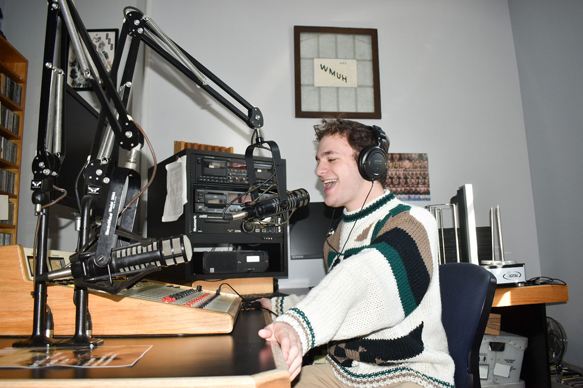 A college student with short brown hair sits in a DJ booth and speaks into a microphone