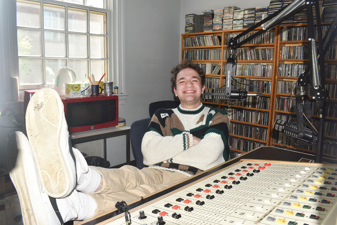 A college student with short brown hair sits in a DJ booth with his feet up near the board and smiles at the camera