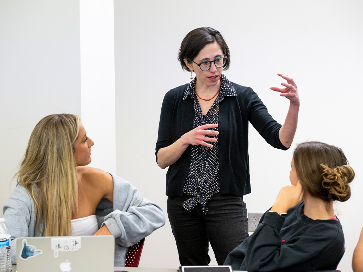 A female professor gesticulates with her hands in a classroom as two students look on.