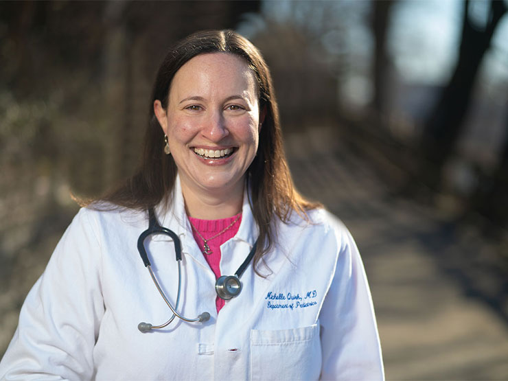 An adult woman smiles at the camera while standing outdoors in a white physician's coat with a stethoscope hanging around her neck.