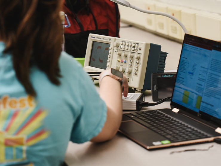 A student's hands reach for a dial on a piece or lab equipment situated near a laptop in a physics lab.