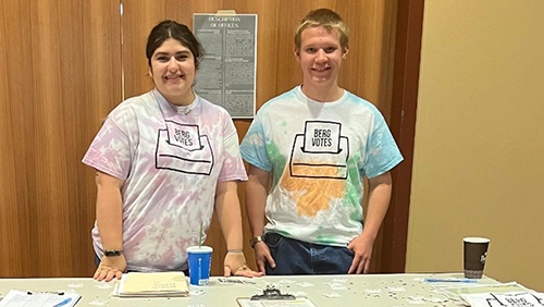 A pair of students in tie-dyed Berg Votes shirts stand at an election registration table.