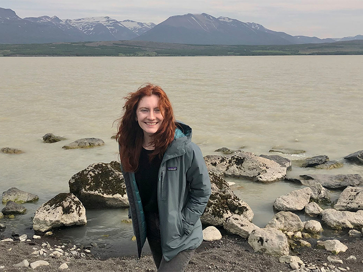 A smiling young adult with long red hair stands in front of rocks, water, and mountains