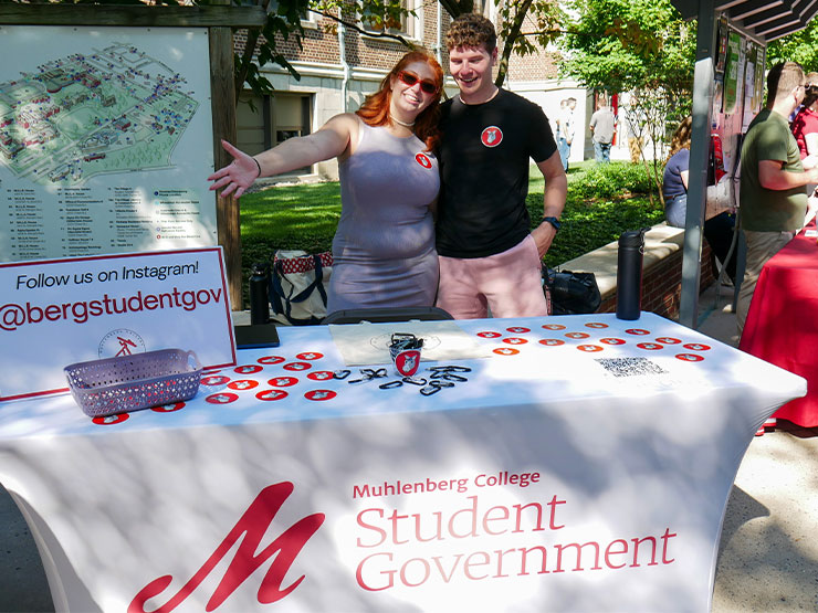 A pair of student stand close to one another, smiling, while tabling for the Muhlenberg Student Government Association on a sunny day.
