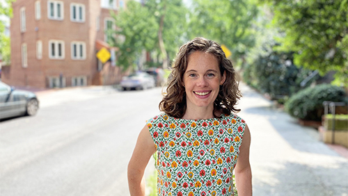 A white woman with curly brown hair wearing a floral dress stands on a city street