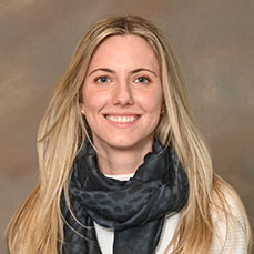 Woman with blonde hair wearing dark grey scarf posing for faculty member portrait in front of abstract background.