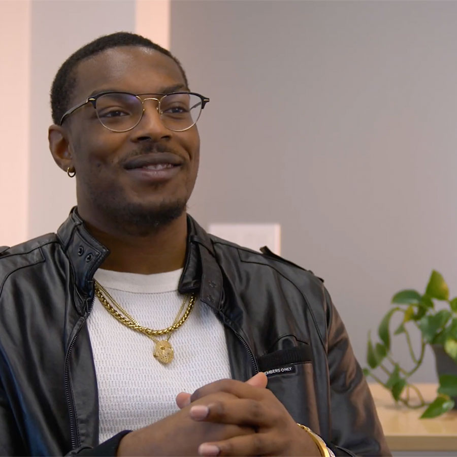 A young adult in a black leather jacket, white shirt, gold chain and glasses speaks to the camera.