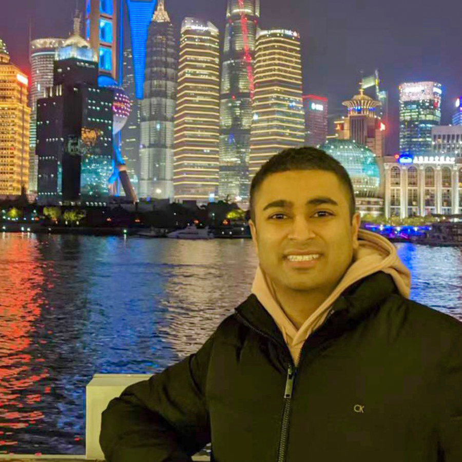 A man in a black jacket leans against a railing along a river with the brilliant lights of a nighttime city skyline in the distance.