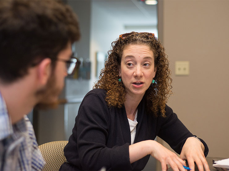 An adult with curly shoulder-length hair speaks with a young adult at a table.