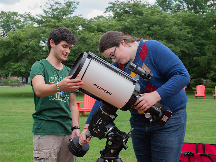 A pair of students adjust a bulky white telescope in a clearing surrounded by forest.