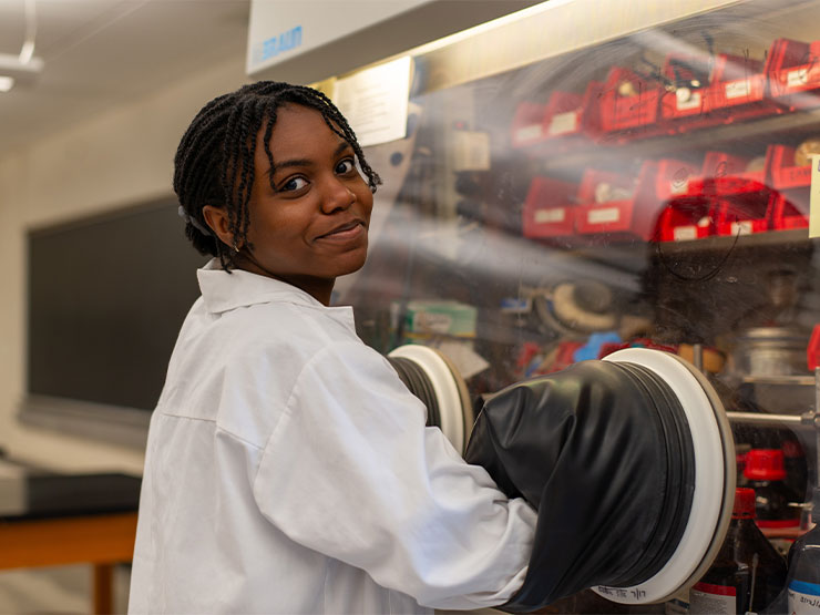 A student, wearing a white lab coat, turns to grin at the camera