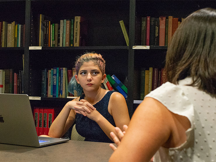 A student with colorful hair sits in front of an open laptop with bookshelves on the wall behind and listens to an instructor speak.
