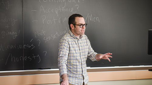 An instructor speaks to a class while standing in front of a chalkboard.