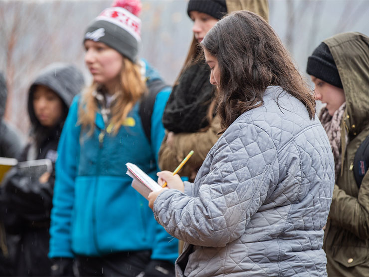 Groups of students in hats and jackets take notes while standing outdoors in soggy weather.