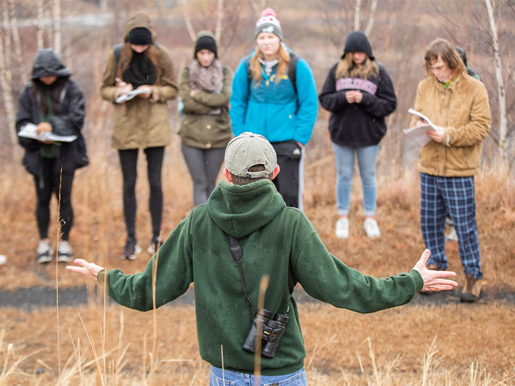 A group of students take notes while someone speaks with them, binoculars around his shoulder, outdoors.