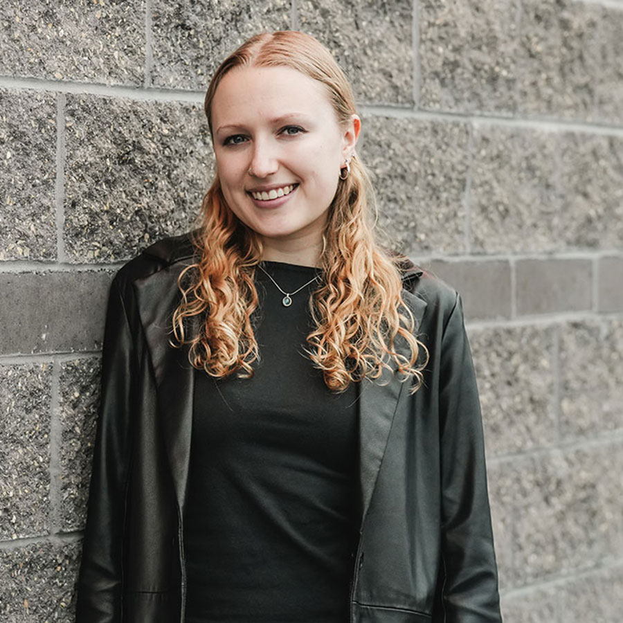 Daisy Cunningham, smiling, stands against a grey brick wall.