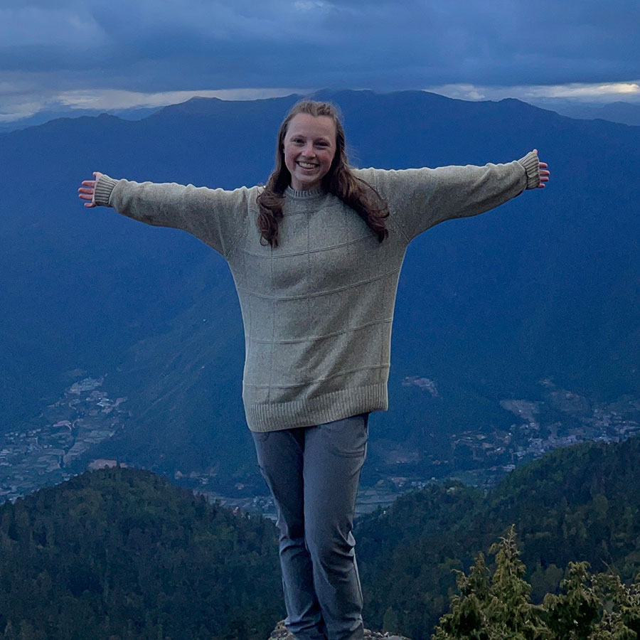 A young adult woman stands, arms outstretched, on a high elevation lookout, with blue mountains stretching into the distance.