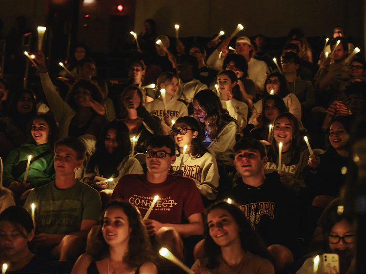 An auditorium of student faces are lit by soft electronic candlelight during a ceremony for first-year students.