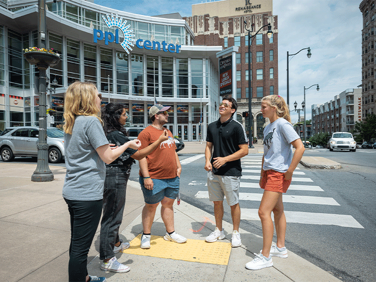 A group of young adults speak to one another at a busy downtown intersection with a sporting arena in the background.