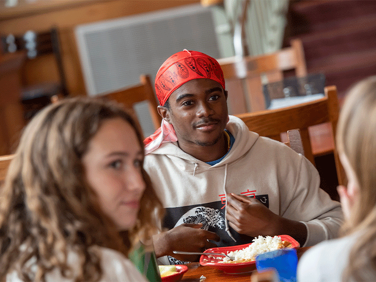 A young adult wearing a white sweatshirt and red bandana on his head looks across a dining hall table at a group of other young adults.