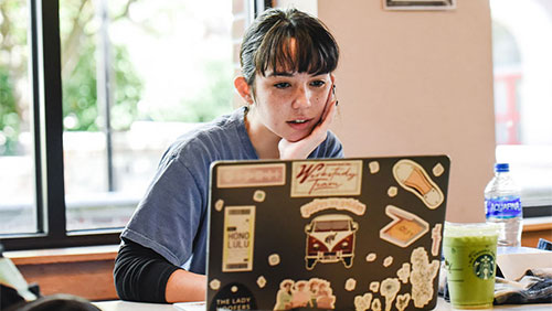 A young adult, resting their chin in their palm, looks at an open laptop.