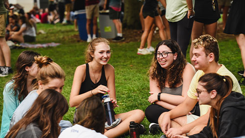 A group of young adults laugh and chat while sitting on a college lawn.