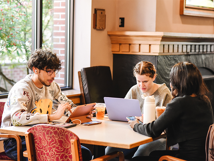 A group of three students work on their laptops and tablets around a table cluttered with notebooks, coffee cups and other beverages.