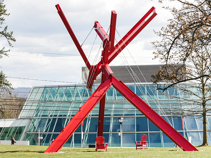 A large, red modern art sculpture stands on the grass with red chairs below and a glass structure situated behind it.