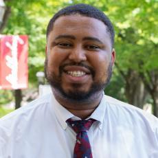 A person in a white dress shirt and tie smiles a the camera with a Muhlenberg flag in the distance.