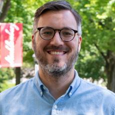 A person with glasses and a blue dress shirt smiles at the camera with a Muhlenberg flag in the distance.