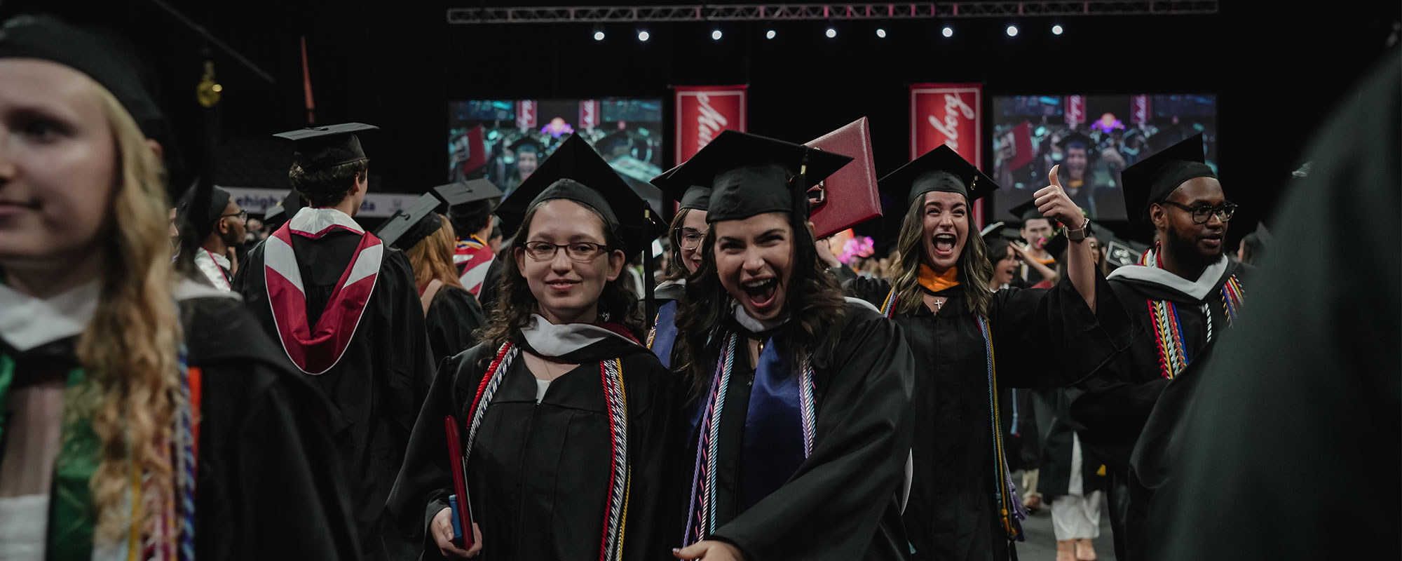 Recent graduates offer wide smiles and exclamations while walking in a procession during a commencement ceremony.