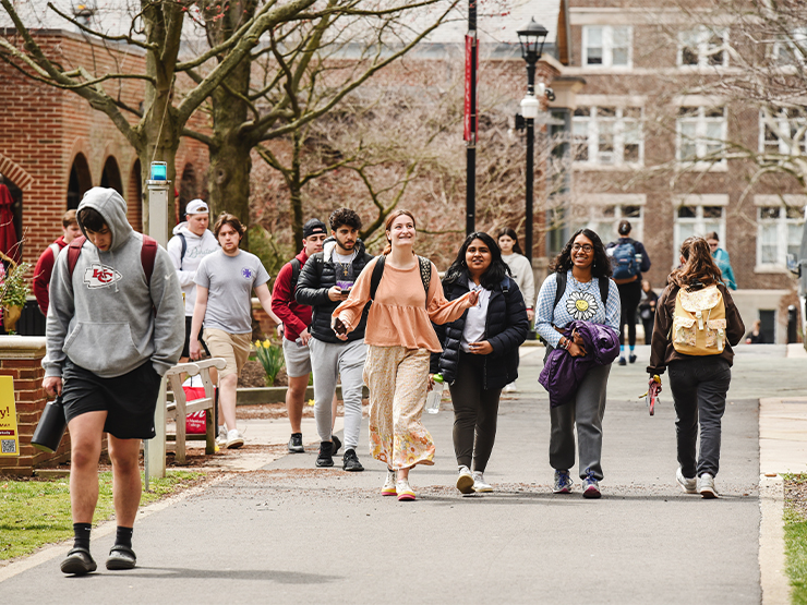 Students fill an walkway on a college campus surrounded by newly budding spring trees.