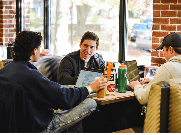 Three students chat while sitting around a table in a cafe area, the table in front of them cluttered with water bottles and laptops.