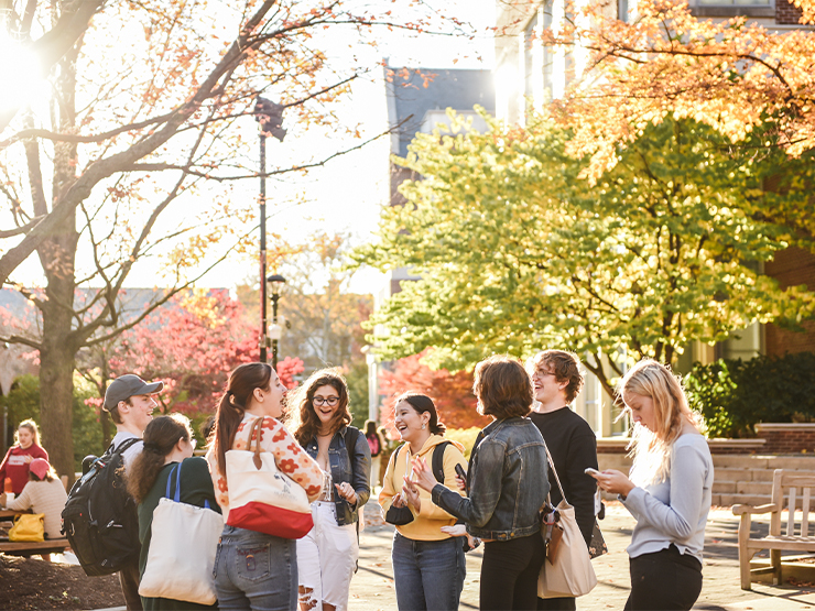 A group of students gather and laugh in a plaza on a college campus, surrounded by colorful fall foliage.