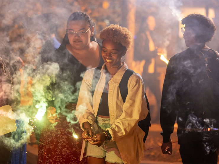 Students gather at night on a College campus holding bright-colored sparklers.